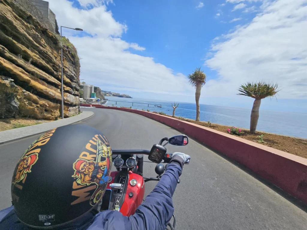 Cruising with Danny and Peter, Holland America Line, Nieuw Statendam, Funchal, Madeira. Portugal, History Adventure | A vibrant collage capturing a day in Funchal, Madeira: a sleek trike navigates winding roads with lush green hills in the background; the panoramic view from Pico dos Barcelos showcases Funchal Bay and the Desertas Islands under a clear sky; Cascata dos Anjos waterfall cascades from cliffs onto a road, surrounded by verdant greenery; Praia da Madalena do Mar’s pebbled beach and black volcanic sand meet gentle waves with towering cliffs above; the misty Valley of the Nuns nestles between rugged mountains, dotted with stone houses and chestnut groves; Cabo Girão’s glass-bottomed skywalk reveals a dizzying drop to the Atlantic and terraced fields below; a plate of warm bolo do caco flatbread filled with steak sits on a café table in Funchal’s colorful old town; the CR7 Museum displays Cristiano Ronaldo’s trophies and jerseys with a waterfront view of the Nieuw Statendam; and Nami Sushi Bar’s elegant sushi platter gleams under soft lighting aboard the ship.