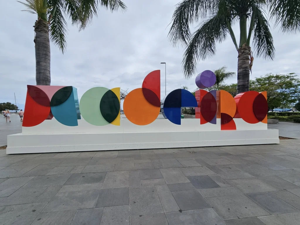 Cruising with Danny and Peter, Holland America Line, Nieuw Statendam, Funchal, Madeira. Portugal, History Adventure | A vibrant collage capturing a day in Funchal, Madeira: a sleek trike navigates winding roads with lush green hills in the background; the panoramic view from Pico dos Barcelos showcases Funchal Bay and the Desertas Islands under a clear sky; Cascata dos Anjos waterfall cascades from cliffs onto a road, surrounded by verdant greenery; Praia da Madalena do Mar’s pebbled beach and black volcanic sand meet gentle waves with towering cliffs above; the misty Valley of the Nuns nestles between rugged mountains, dotted with stone houses and chestnut groves; Cabo Girão’s glass-bottomed skywalk reveals a dizzying drop to the Atlantic and terraced fields below; a plate of warm bolo do caco flatbread filled with steak sits on a café table in Funchal’s colorful old town; the CR7 Museum displays Cristiano Ronaldo’s trophies and jerseys with a waterfront view of the Nieuw Statendam; and Nami Sushi Bar’s elegant sushi platter gleams under soft lighting aboard the ship.