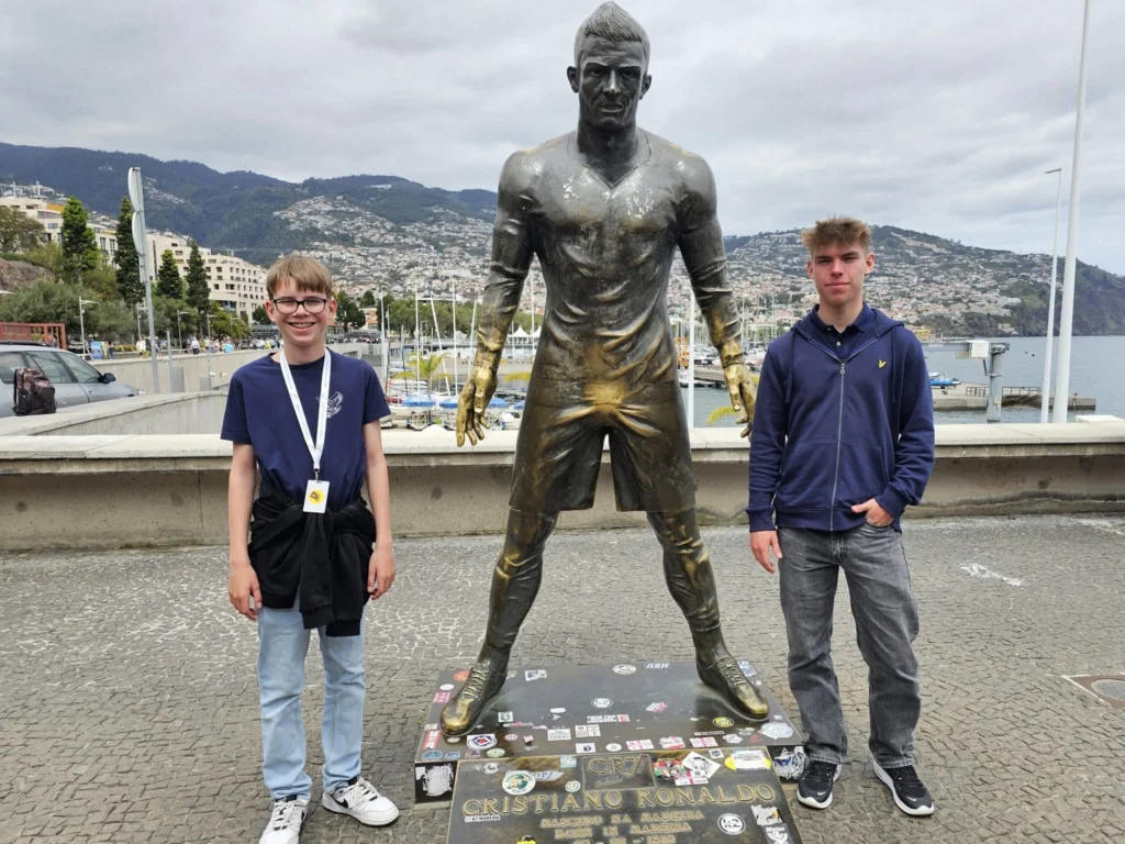 Cruising with Danny and Peter, Holland America Line, Nieuw Statendam, Funchal, Madeira. Portugal, History Adventure | A vibrant collage capturing a day in Funchal, Madeira: a sleek trike navigates winding roads with lush green hills in the background; the panoramic view from Pico dos Barcelos showcases Funchal Bay and the Desertas Islands under a clear sky; Cascata dos Anjos waterfall cascades from cliffs onto a road, surrounded by verdant greenery; Praia da Madalena do Mar’s pebbled beach and black volcanic sand meet gentle waves with towering cliffs above; the misty Valley of the Nuns nestles between rugged mountains, dotted with stone houses and chestnut groves; Cabo Girão’s glass-bottomed skywalk reveals a dizzying drop to the Atlantic and terraced fields below; a plate of warm bolo do caco flatbread filled with steak sits on a café table in Funchal’s colorful old town; the CR7 Museum displays Cristiano Ronaldo’s trophies and jerseys with a waterfront view of the Nieuw Statendam; and Nami Sushi Bar’s elegant sushi platter gleams under soft lighting aboard the ship.