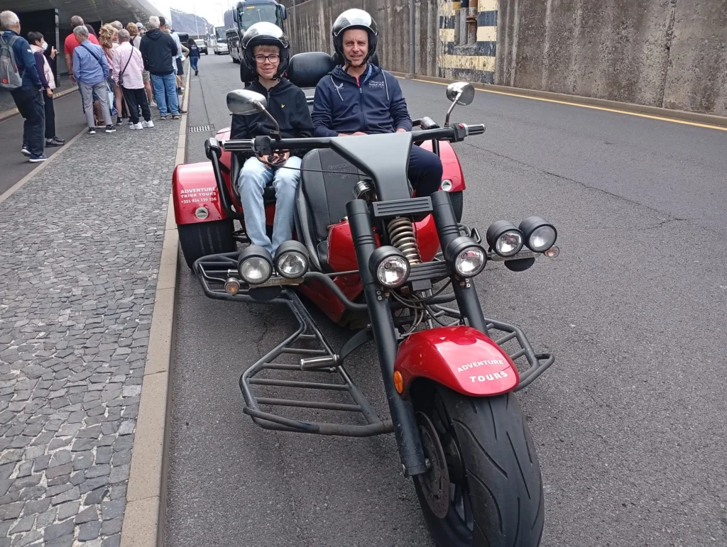 Cruising with Danny and Peter, Holland America Line, Nieuw Statendam, Funchal, Madeira. Portugal, History Adventure | A vibrant collage capturing a day in Funchal, Madeira: a sleek trike navigates winding roads with lush green hills in the background; the panoramic view from Pico dos Barcelos showcases Funchal Bay and the Desertas Islands under a clear sky; Cascata dos Anjos waterfall cascades from cliffs onto a road, surrounded by verdant greenery; Praia da Madalena do Mar’s pebbled beach and black volcanic sand meet gentle waves with towering cliffs above; the misty Valley of the Nuns nestles between rugged mountains, dotted with stone houses and chestnut groves; Cabo Girão’s glass-bottomed skywalk reveals a dizzying drop to the Atlantic and terraced fields below; a plate of warm bolo do caco flatbread filled with steak sits on a café table in Funchal’s colorful old town; the CR7 Museum displays Cristiano Ronaldo’s trophies and jerseys with a waterfront view of the Nieuw Statendam; and Nami Sushi Bar’s elegant sushi platter gleams under soft lighting aboard the ship.