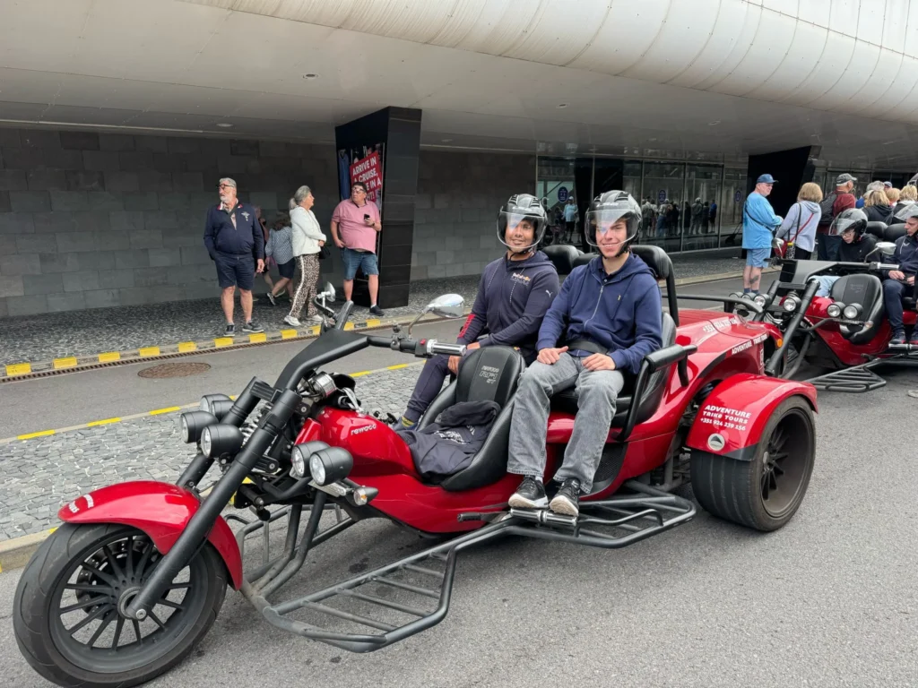 Cruising with Danny and Peter, Holland America Line, Nieuw Statendam, Funchal, Madeira. Portugal, History Adventure | A vibrant collage capturing a day in Funchal, Madeira: a sleek trike navigates winding roads with lush green hills in the background; the panoramic view from Pico dos Barcelos showcases Funchal Bay and the Desertas Islands under a clear sky; Cascata dos Anjos waterfall cascades from cliffs onto a road, surrounded by verdant greenery; Praia da Madalena do Mar’s pebbled beach and black volcanic sand meet gentle waves with towering cliffs above; the misty Valley of the Nuns nestles between rugged mountains, dotted with stone houses and chestnut groves; Cabo Girão’s glass-bottomed skywalk reveals a dizzying drop to the Atlantic and terraced fields below; a plate of warm bolo do caco flatbread filled with steak sits on a café table in Funchal’s colorful old town; the CR7 Museum displays Cristiano Ronaldo’s trophies and jerseys with a waterfront view of the Nieuw Statendam; and Nami Sushi Bar’s elegant sushi platter gleams under soft lighting aboard the ship.