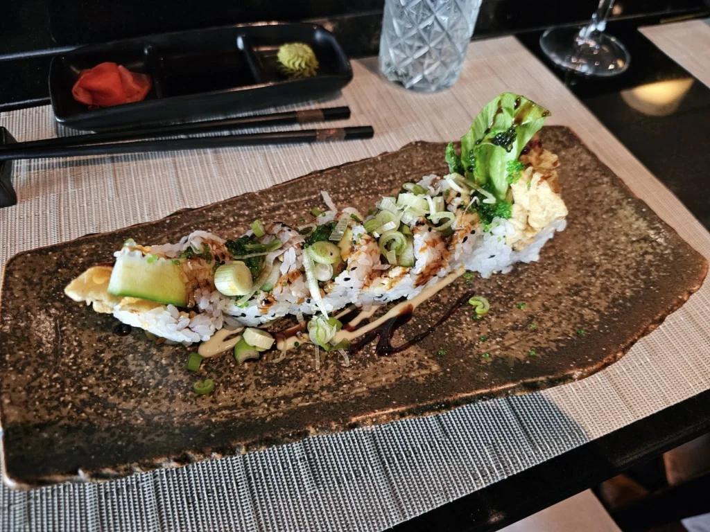 Cruising with Danny and Peter, Holland America Line, Nieuw Statendam, Funchal, Madeira. Portugal, History Adventure | A vibrant collage capturing a day in Funchal, Madeira: a sleek trike navigates winding roads with lush green hills in the background; the panoramic view from Pico dos Barcelos showcases Funchal Bay and the Desertas Islands under a clear sky; Cascata dos Anjos waterfall cascades from cliffs onto a road, surrounded by verdant greenery; Praia da Madalena do Mar’s pebbled beach and black volcanic sand meet gentle waves with towering cliffs above; the misty Valley of the Nuns nestles between rugged mountains, dotted with stone houses and chestnut groves; Cabo Girão’s glass-bottomed skywalk reveals a dizzying drop to the Atlantic and terraced fields below; a plate of warm bolo do caco flatbread filled with steak sits on a café table in Funchal’s colorful old town; the CR7 Museum displays Cristiano Ronaldo’s trophies and jerseys with a waterfront view of the Nieuw Statendam; and Nami Sushi Bar’s elegant sushi platter gleams under soft lighting aboard the ship.