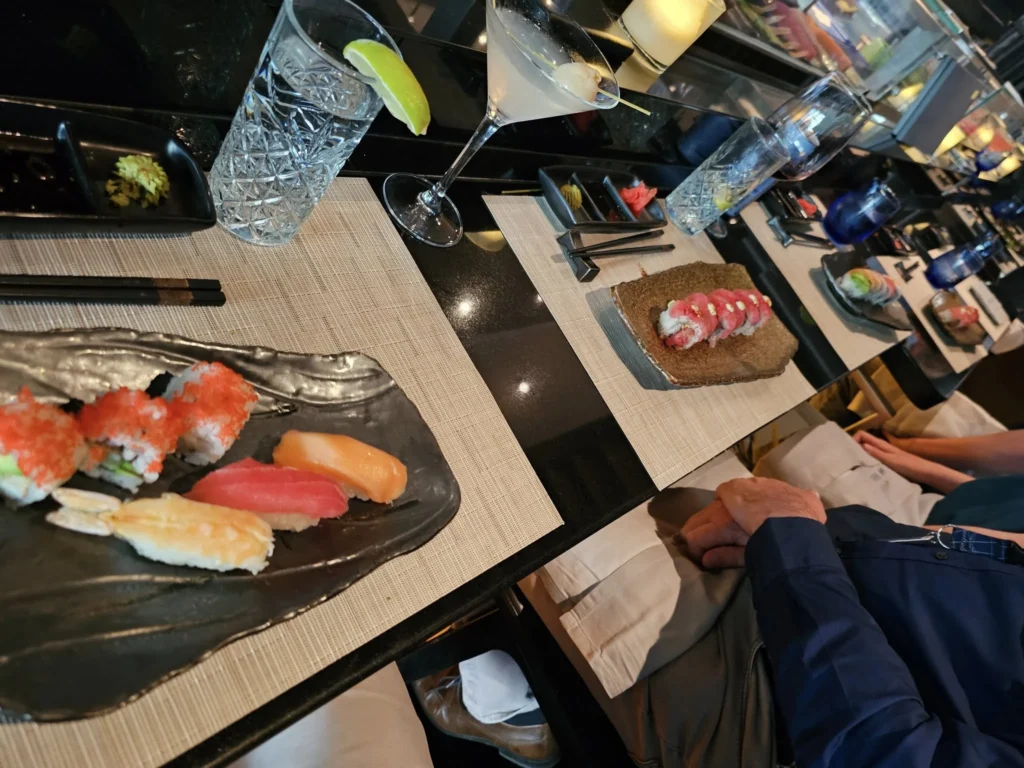 Cruising with Danny and Peter, Holland America Line, Nieuw Statendam, Funchal, Madeira. Portugal, History Adventure | A vibrant collage capturing a day in Funchal, Madeira: a sleek trike navigates winding roads with lush green hills in the background; the panoramic view from Pico dos Barcelos showcases Funchal Bay and the Desertas Islands under a clear sky; Cascata dos Anjos waterfall cascades from cliffs onto a road, surrounded by verdant greenery; Praia da Madalena do Mar’s pebbled beach and black volcanic sand meet gentle waves with towering cliffs above; the misty Valley of the Nuns nestles between rugged mountains, dotted with stone houses and chestnut groves; Cabo Girão’s glass-bottomed skywalk reveals a dizzying drop to the Atlantic and terraced fields below; a plate of warm bolo do caco flatbread filled with steak sits on a café table in Funchal’s colorful old town; the CR7 Museum displays Cristiano Ronaldo’s trophies and jerseys with a waterfront view of the Nieuw Statendam; and Nami Sushi Bar’s elegant sushi platter gleams under soft lighting aboard the ship.