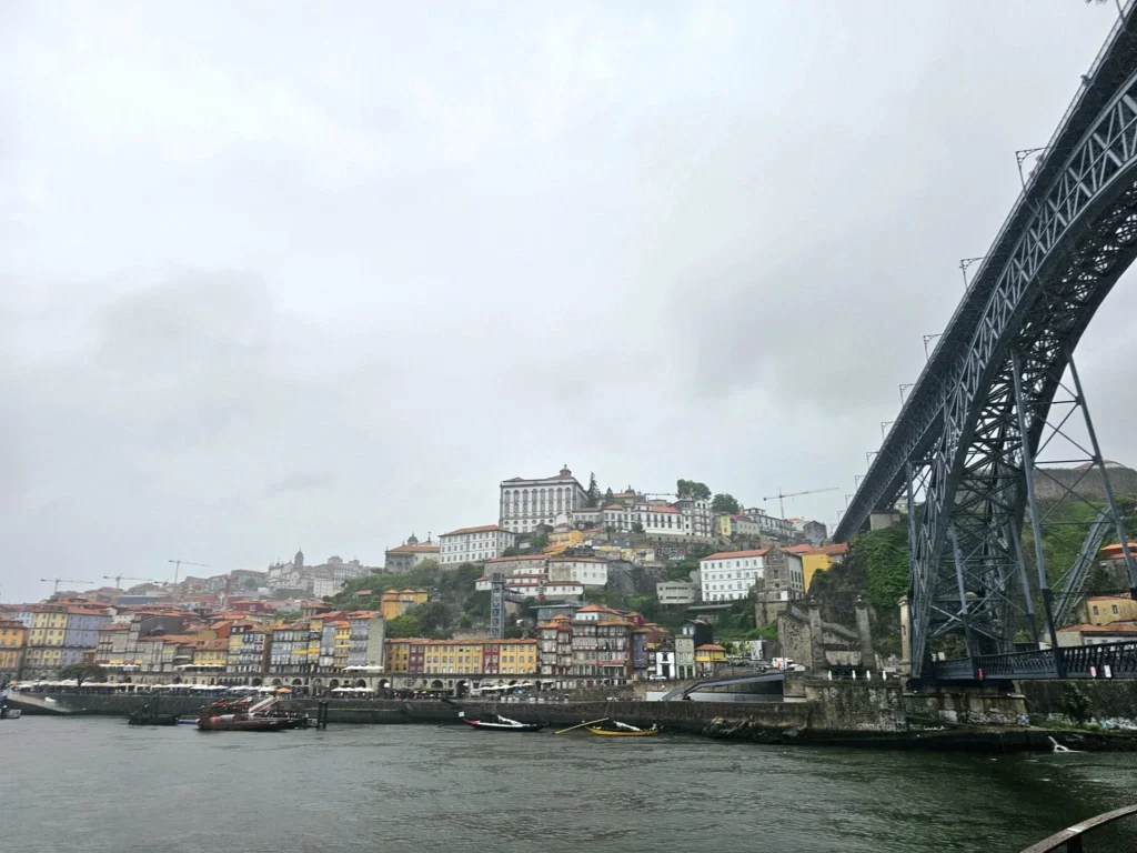 Cruising with Danny and Peter, Holland America Line, Nieuw Statendam, Leixoes, Porto, Portugal, Rainy Adventure | A vibrant collage capturing a rainy day adventure in Porto, Portugal, from the Holland America Line’s Nieuw Statendam cruise. The scene includes a colorful tuk-tuk navigating Porto’s wet, cobblestone streets with historic buildings in the background, the iconic Dom Luís I Bridge spanning the Douro River under gray skies, the majestic Porto Cathedral with its Gothic facade glistening in the rain, and the intricate blue-and-white azulejo tiles of São Bento Station’s atrium. Onboard, a beautifully plated dish from Tamarind restaurant showcases exotic flavors, while the lively Lido Fun Fair on the Lido Midship deck features children enjoying carnival games under a retractable roof, with colorful decorations and cotton candy adding festive cheer.