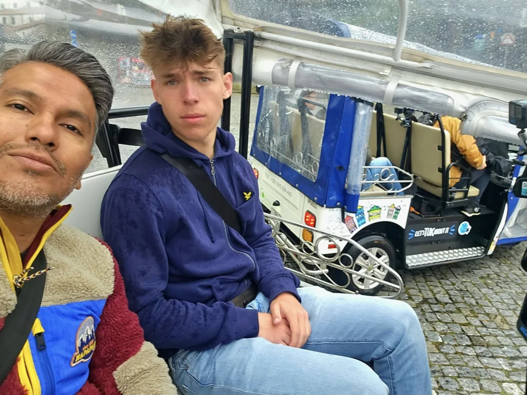 Cruising with Danny and Peter, Holland America Line, Nieuw Statendam, Leixoes, Porto, Portugal, Rainy Adventure | A vibrant collage capturing a rainy day adventure in Porto, Portugal, from the Holland America Line’s Nieuw Statendam cruise. The scene includes a colorful tuk-tuk navigating Porto’s wet, cobblestone streets with historic buildings in the background, the iconic Dom Luís I Bridge spanning the Douro River under gray skies, the majestic Porto Cathedral with its Gothic facade glistening in the rain, and the intricate blue-and-white azulejo tiles of São Bento Station’s atrium. Onboard, a beautifully plated dish from Tamarind restaurant showcases exotic flavors, while the lively Lido Fun Fair on the Lido Midship deck features children enjoying carnival games under a retractable roof, with colorful decorations and cotton candy adding festive cheer.