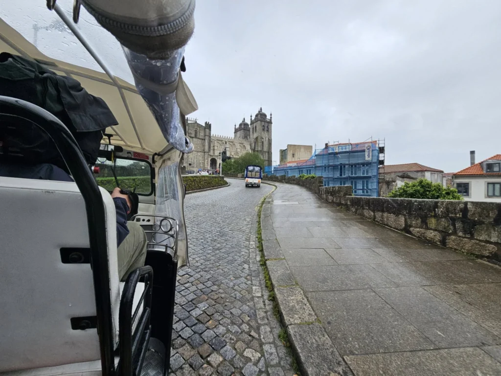 Cruising with Danny and Peter, Holland America Line, Nieuw Statendam, Leixoes, Porto, Portugal, Rainy Adventure | A vibrant collage capturing a rainy day adventure in Porto, Portugal, from the Holland America Line’s Nieuw Statendam cruise. The scene includes a colorful tuk-tuk navigating Porto’s wet, cobblestone streets with historic buildings in the background, the iconic Dom Luís I Bridge spanning the Douro River under gray skies, the majestic Porto Cathedral with its Gothic facade glistening in the rain, and the intricate blue-and-white azulejo tiles of São Bento Station’s atrium. Onboard, a beautifully plated dish from Tamarind restaurant showcases exotic flavors, while the lively Lido Fun Fair on the Lido Midship deck features children enjoying carnival games under a retractable roof, with colorful decorations and cotton candy adding festive cheer.