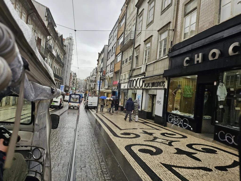Cruising with Danny and Peter, Holland America Line, Nieuw Statendam, Leixoes, Porto, Portugal, Rainy Adventure | A vibrant collage capturing a rainy day adventure in Porto, Portugal, from the Holland America Line’s Nieuw Statendam cruise. The scene includes a colorful tuk-tuk navigating Porto’s wet, cobblestone streets with historic buildings in the background, the iconic Dom Luís I Bridge spanning the Douro River under gray skies, the majestic Porto Cathedral with its Gothic facade glistening in the rain, and the intricate blue-and-white azulejo tiles of São Bento Station’s atrium. Onboard, a beautifully plated dish from Tamarind restaurant showcases exotic flavors, while the lively Lido Fun Fair on the Lido Midship deck features children enjoying carnival games under a retractable roof, with colorful decorations and cotton candy adding festive cheer.