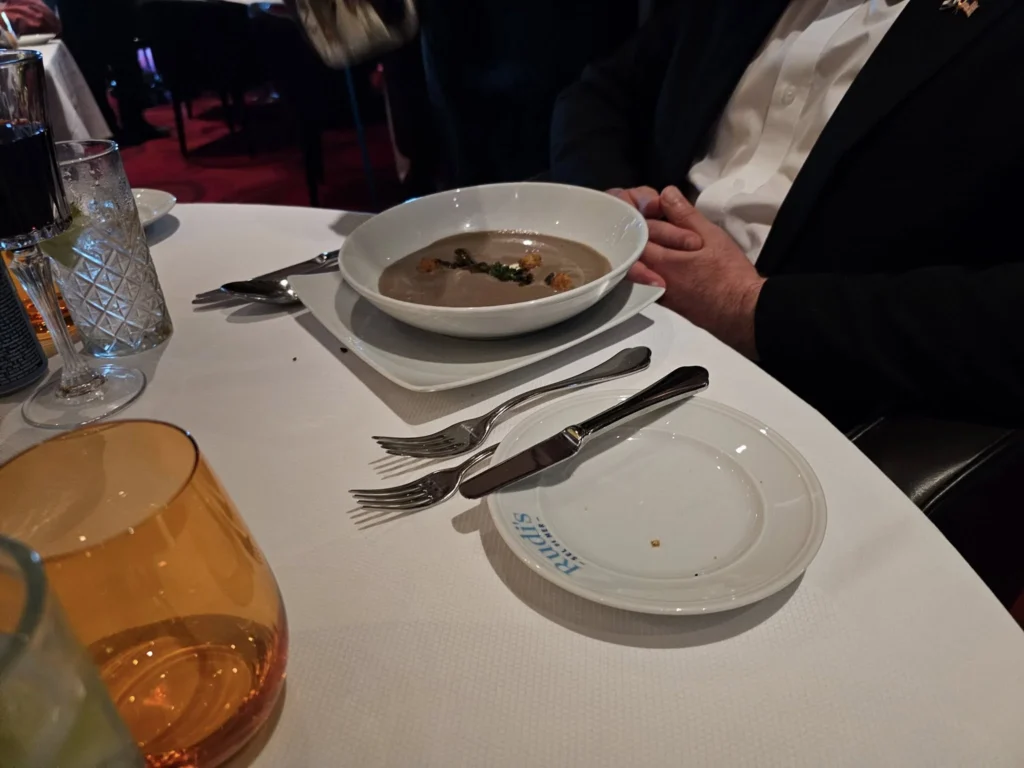 Cruising with Danny and Peter, Holland America Line, Nieuw Statendam, Biscay Bliss Sea Day | A vibrant collage capturing a sea day aboard Holland America Line’s Nieuw Statendam in the Bay of Biscay. The scene includes a pod of dolphins leaping gracefully through shimmering blue waves alongside the ship, their sleek bodies catching the sunlight. Passengers walk energetically on the sunlit deck during a charity event, with the vast ocean horizon stretching behind them. The elegant interior of Rudi’s Sel de Mer restaurant features a beautifully set table with a plate of seared scallops and a chocolate soufflé, illuminated by warm lighting. The World Stage theater glows with a large LED screen displaying Dover’s white cliffs, as the audience listens attentively to a port talk. A couple poses for a photoshoot on deck at sunset, framed by the golden glow of the Bay of Biscay’s horizon, with the ship’s sleek railings in the foreground.