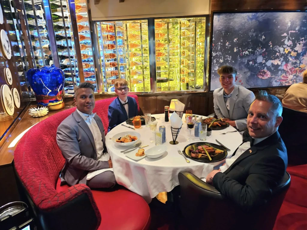 Cruising with Danny and Peter, Holland America Line, Nieuw Statendam, Biscay Bliss Sea Day | A vibrant collage capturing a sea day aboard Holland America Line’s Nieuw Statendam in the Bay of Biscay. The scene includes a pod of dolphins leaping gracefully through shimmering blue waves alongside the ship, their sleek bodies catching the sunlight. Passengers walk energetically on the sunlit deck during a charity event, with the vast ocean horizon stretching behind them. The elegant interior of Rudi’s Sel de Mer restaurant features a beautifully set table with a plate of seared scallops and a chocolate soufflé, illuminated by warm lighting. The World Stage theater glows with a large LED screen displaying Dover’s white cliffs, as the audience listens attentively to a port talk. A couple poses for a photoshoot on deck at sunset, framed by the golden glow of the Bay of Biscay’s horizon, with the ship’s sleek railings in the foreground.