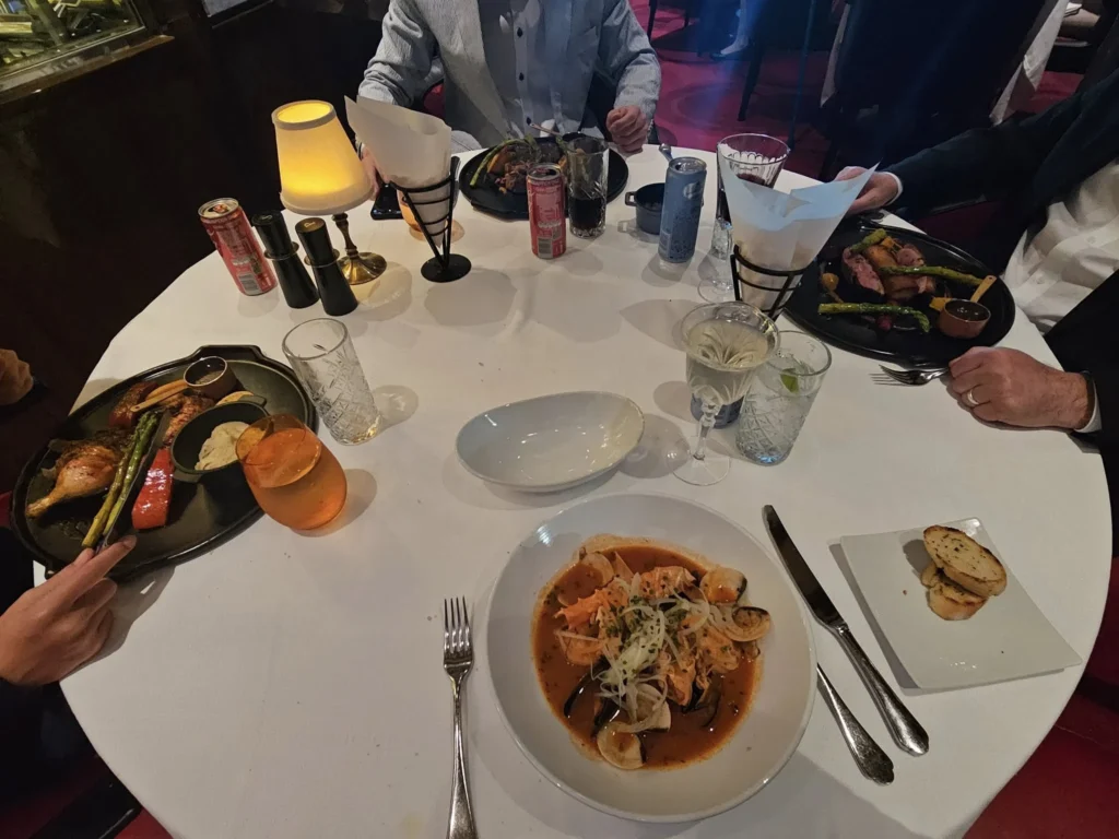 Cruising with Danny and Peter, Holland America Line, Nieuw Statendam, Biscay Bliss Sea Day | A vibrant collage capturing a sea day aboard Holland America Line’s Nieuw Statendam in the Bay of Biscay. The scene includes a pod of dolphins leaping gracefully through shimmering blue waves alongside the ship, their sleek bodies catching the sunlight. Passengers walk energetically on the sunlit deck during a charity event, with the vast ocean horizon stretching behind them. The elegant interior of Rudi’s Sel de Mer restaurant features a beautifully set table with a plate of seared scallops and a chocolate soufflé, illuminated by warm lighting. The World Stage theater glows with a large LED screen displaying Dover’s white cliffs, as the audience listens attentively to a port talk. A couple poses for a photoshoot on deck at sunset, framed by the golden glow of the Bay of Biscay’s horizon, with the ship’s sleek railings in the foreground.