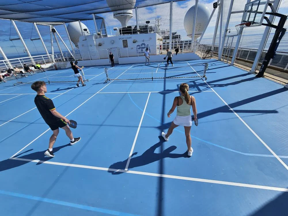 Cruising with Danny and Peter, Holland America Line, Nieuw Statendam, English Channel Sea Day | A montage of a sea day on Holland America’s Nieuw Statendam sailing the English Channel: calm waters meet Dover’s distant white cliffs from a balcony; Sven and Liam play pickleball on a sunlit deck with cheering crowds and a blue sea backdrop; vibrant cakes—chocolate, fruit, and cream-topped—fill the Lido Market as crew serve slices; a candlelit Canaletto table holds Italian dishes in a cozy, rustic dining room.