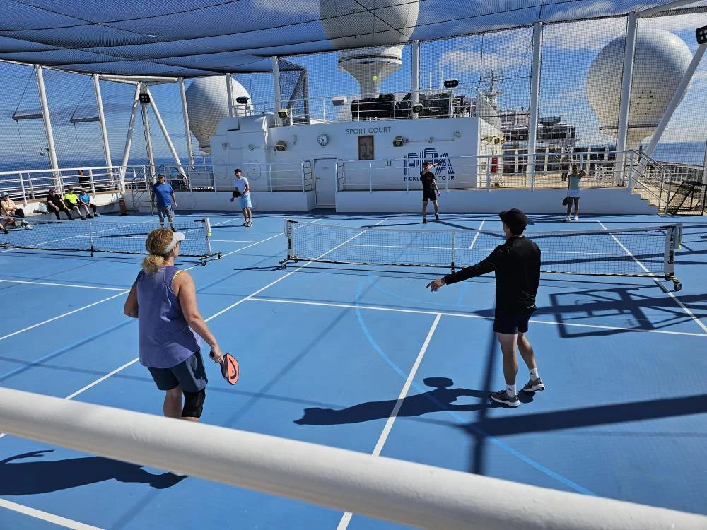 Cruising with Danny and Peter, Holland America Line, Nieuw Statendam, English Channel Sea Day | A montage of a sea day on Holland America’s Nieuw Statendam sailing the English Channel: calm waters meet Dover’s distant white cliffs from a balcony; Sven and Liam play pickleball on a sunlit deck with cheering crowds and a blue sea backdrop; vibrant cakes—chocolate, fruit, and cream-topped—fill the Lido Market as crew serve slices; a candlelit Canaletto table holds Italian dishes in a cozy, rustic dining room.