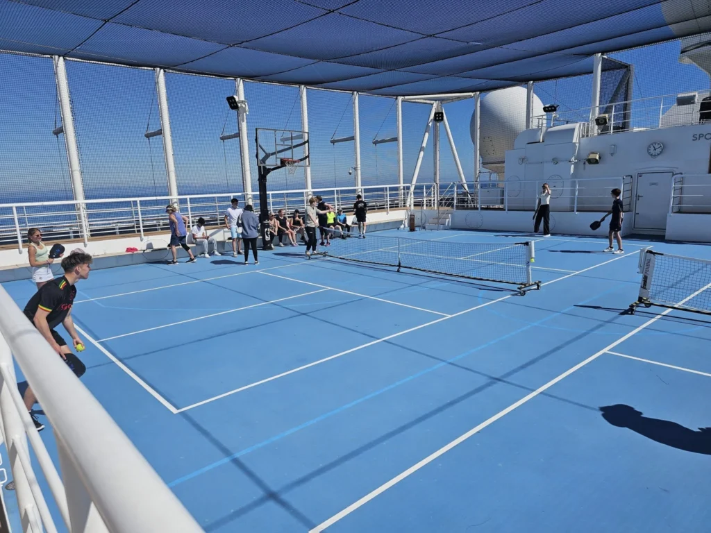 Cruising with Danny and Peter, Holland America Line, Nieuw Statendam, English Channel Sea Day | A montage of a sea day on Holland America’s Nieuw Statendam sailing the English Channel: calm waters meet Dover’s distant white cliffs from a balcony; Sven and Liam play pickleball on a sunlit deck with cheering crowds and a blue sea backdrop; vibrant cakes—chocolate, fruit, and cream-topped—fill the Lido Market as crew serve slices; a candlelit Canaletto table holds Italian dishes in a cozy, rustic dining room.