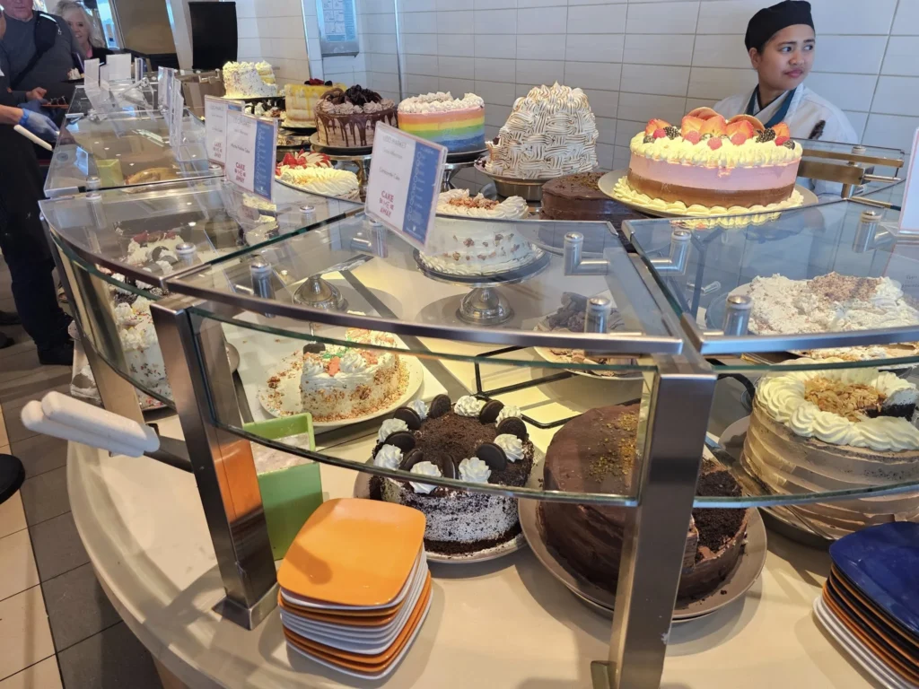 Cruising with Danny and Peter, Holland America Line, Nieuw Statendam, English Channel Sea Day | A montage of a sea day on Holland America’s Nieuw Statendam sailing the English Channel: calm waters meet Dover’s distant white cliffs from a balcony; Sven and Liam play pickleball on a sunlit deck with cheering crowds and a blue sea backdrop; vibrant cakes—chocolate, fruit, and cream-topped—fill the Lido Market as crew serve slices; a candlelit Canaletto table holds Italian dishes in a cozy, rustic dining room.