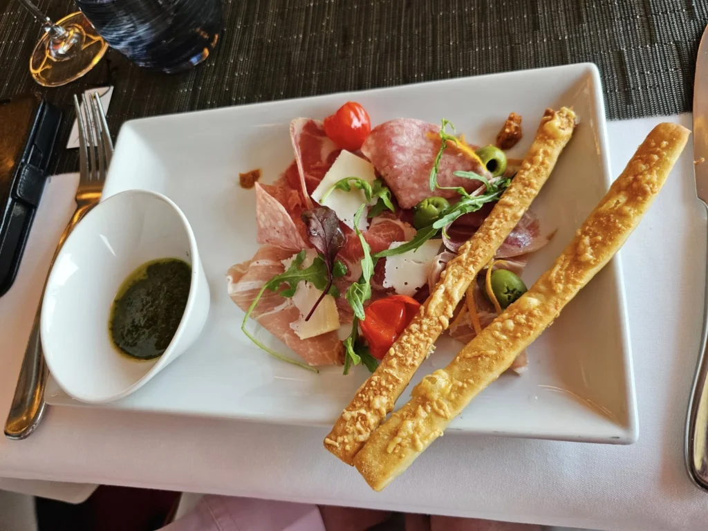 Cruising with Danny and Peter, Holland America Line, Nieuw Statendam, English Channel Sea Day | A montage of a sea day on Holland America’s Nieuw Statendam sailing the English Channel: calm waters meet Dover’s distant white cliffs from a balcony; Sven and Liam play pickleball on a sunlit deck with cheering crowds and a blue sea backdrop; vibrant cakes—chocolate, fruit, and cream-topped—fill the Lido Market as crew serve slices; a candlelit Canaletto table holds Italian dishes in a cozy, rustic dining room.