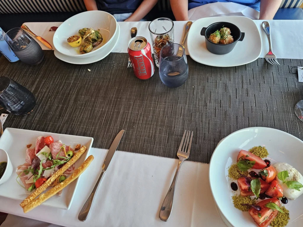 Cruising with Danny and Peter, Holland America Line, Nieuw Statendam, English Channel Sea Day | A montage of a sea day on Holland America’s Nieuw Statendam sailing the English Channel: calm waters meet Dover’s distant white cliffs from a balcony; Sven and Liam play pickleball on a sunlit deck with cheering crowds and a blue sea backdrop; vibrant cakes—chocolate, fruit, and cream-topped—fill the Lido Market as crew serve slices; a candlelit Canaletto table holds Italian dishes in a cozy, rustic dining room.