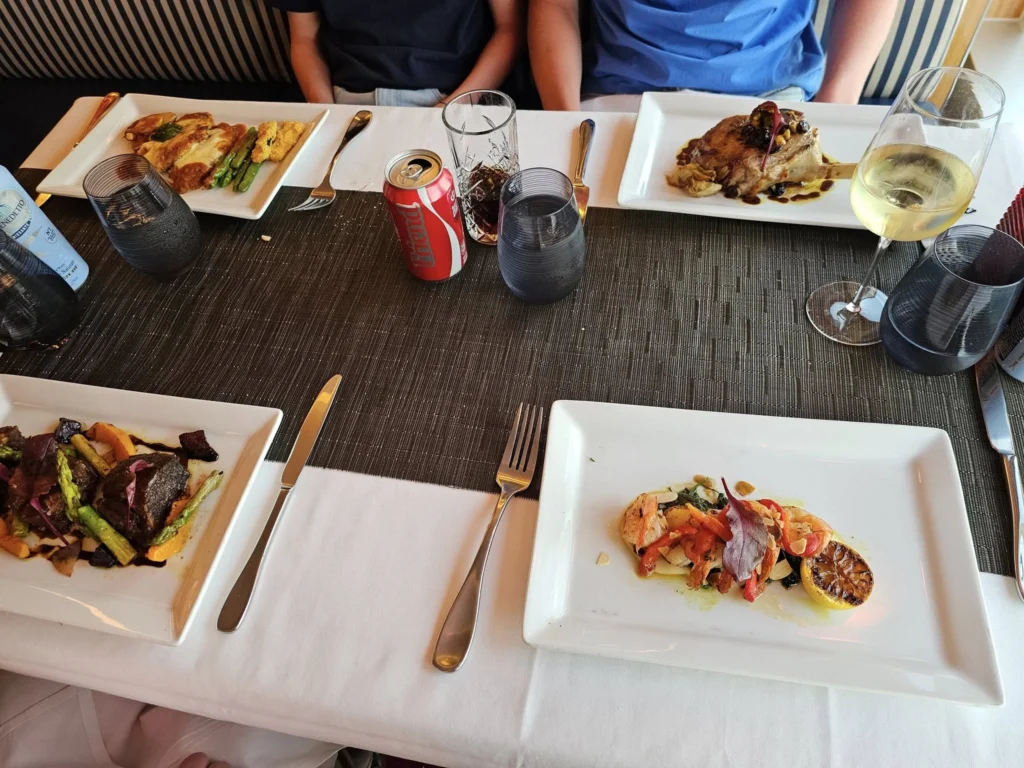 Cruising with Danny and Peter, Holland America Line, Nieuw Statendam, English Channel Sea Day | A montage of a sea day on Holland America’s Nieuw Statendam sailing the English Channel: calm waters meet Dover’s distant white cliffs from a balcony; Sven and Liam play pickleball on a sunlit deck with cheering crowds and a blue sea backdrop; vibrant cakes—chocolate, fruit, and cream-topped—fill the Lido Market as crew serve slices; a candlelit Canaletto table holds Italian dishes in a cozy, rustic dining room.