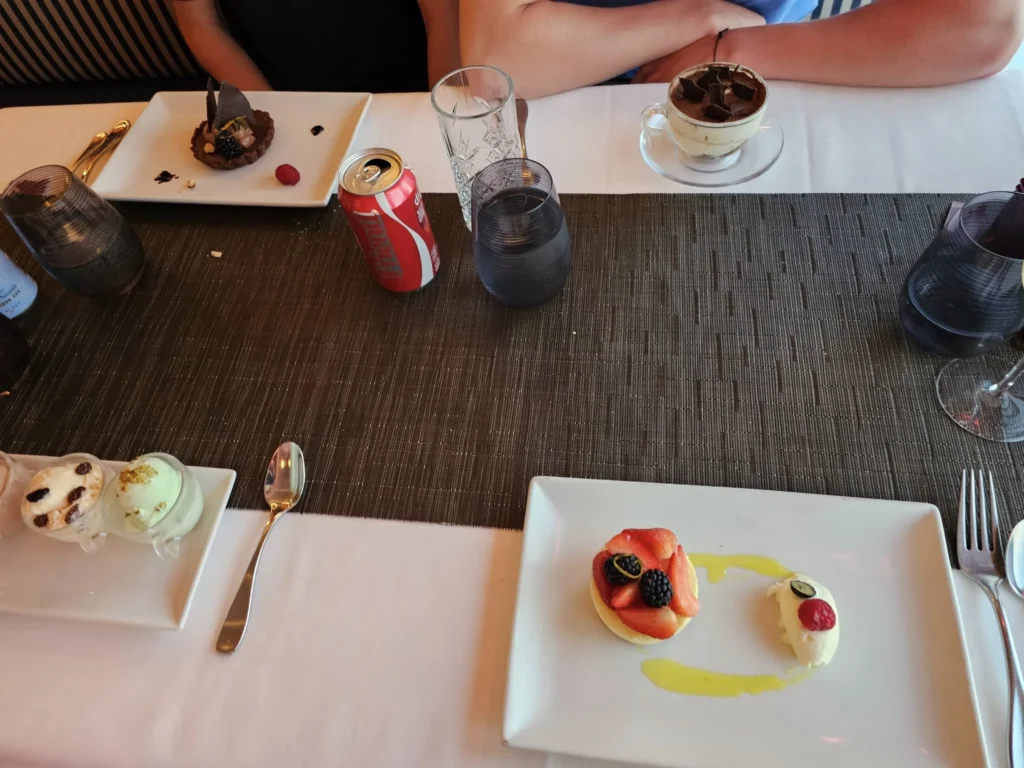 Cruising with Danny and Peter, Holland America Line, Nieuw Statendam, English Channel Sea Day | A montage of a sea day on Holland America’s Nieuw Statendam sailing the English Channel: calm waters meet Dover’s distant white cliffs from a balcony; Sven and Liam play pickleball on a sunlit deck with cheering crowds and a blue sea backdrop; vibrant cakes—chocolate, fruit, and cream-topped—fill the Lido Market as crew serve slices; a candlelit Canaletto table holds Italian dishes in a cozy, rustic dining room.