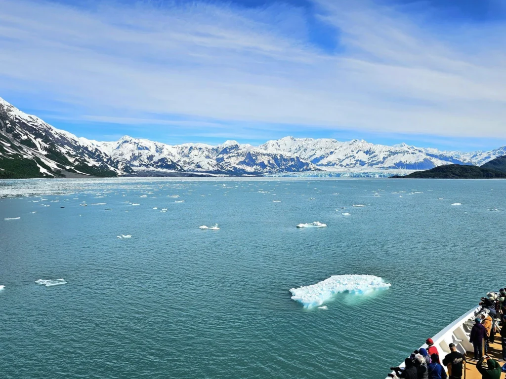 Cruising with Danny and Peter, Holland America Line, Westerdam, Hubbard Glacier, Alaska, United States | View of Hubbard Glacier from the boat during an unforgettable tour, showcasing stunning blue ice and calm Arctic waters. Thrilling ice calving event captures large chunks of ice breaking off and crashing into the water. Mirror-like reflection of the glacier on still waters highlights its majestic blue hues. A delightful picnic with the glacier in the background combines delicious food with breathtaking views, while a close-up of the glacier's intricate ice formations shows deep crevasses and jagged edges.