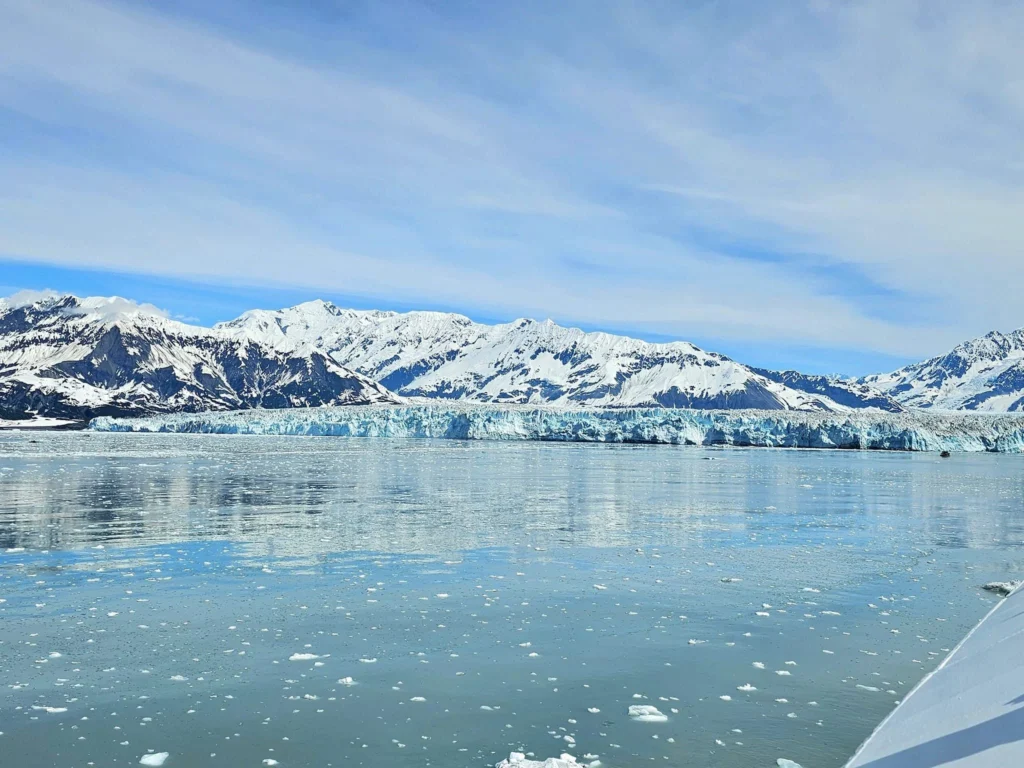 Cruising with Danny and Peter, Holland America Line, Westerdam, Hubbard Glacier, Alaska, United States | View of Hubbard Glacier from the boat during an unforgettable tour, showcasing stunning blue ice and calm Arctic waters. Thrilling ice calving event captures large chunks of ice breaking off and crashing into the water. Mirror-like reflection of the glacier on still waters highlights its majestic blue hues. A delightful picnic with the glacier in the background combines delicious food with breathtaking views, while a close-up of the glacier's intricate ice formations shows deep crevasses and jagged edges.