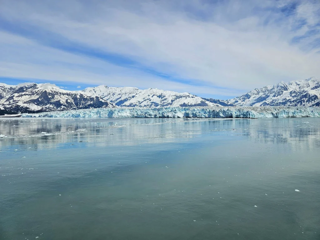 Cruising with Danny and Peter, Holland America Line, Westerdam, Hubbard Glacier, Alaska, United States | View of Hubbard Glacier from the boat during an unforgettable tour, showcasing stunning blue ice and calm Arctic waters. Thrilling ice calving event captures large chunks of ice breaking off and crashing into the water. Mirror-like reflection of the glacier on still waters highlights its majestic blue hues. A delightful picnic with the glacier in the background combines delicious food with breathtaking views, while a close-up of the glacier's intricate ice formations shows deep crevasses and jagged edges.