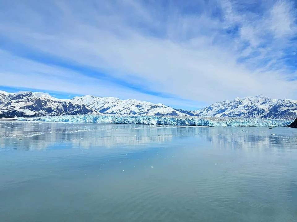 Cruising with Danny and Peter, Holland America Line, Westerdam, Hubbard Glacier, Alaska, United States | View of Hubbard Glacier from the boat during an unforgettable tour, showcasing stunning blue ice and calm Arctic waters. Thrilling ice calving event captures large chunks of ice breaking off and crashing into the water. Mirror-like reflection of the glacier on still waters highlights its majestic blue hues. A delightful picnic with the glacier in the background combines delicious food with breathtaking views, while a close-up of the glacier's intricate ice formations shows deep crevasses and jagged edges.
