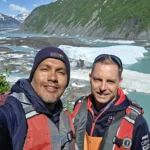 Cruising with Danny and Peter, Holland America Line, Westerdam, Prince William Sound, Valdez, Alaska, United States | A serene panorama of Prince William Sound at dawn, with the luxurious Westerdam cruise ship gliding through calm waters, surrounded by snow-capped mountains and glowing glaciers under a soft pink sky. Humpback whales breach the surface, their tails splashing against the reflective water, while sea otters float playfully in kelp beds and a bald eagle soars overhead. In Valdez Glacier Lake, vibrant blue icebergs drift peacefully, with kayakers in colorful kayaks paddling through the icy maze, dwarfed by the towering Valdez Glacier’s rugged ice face in the background. The Westerdam’s elegant deck features passengers enjoying a gourmet breakfast with panoramic views of the Alaskan wilderness, capturing the harmonious blend of luxury and nature.