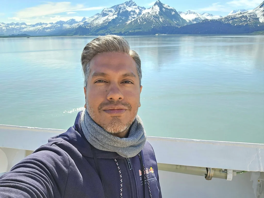 Cruising with Danny and Peter, Holland America Line, Westerdam, Prince William Sound, Valdez, Alaska, United States | A serene panorama of Prince William Sound at dawn, with the luxurious Westerdam cruise ship gliding through calm waters, surrounded by snow-capped mountains and glowing glaciers under a soft pink sky. Humpback whales breach the surface, their tails splashing against the reflective water, while sea otters float playfully in kelp beds and a bald eagle soars overhead. In Valdez Glacier Lake, vibrant blue icebergs drift peacefully, with kayakers in colorful kayaks paddling through the icy maze, dwarfed by the towering Valdez Glacier’s rugged ice face in the background. The Westerdam’s elegant deck features passengers enjoying a gourmet breakfast with panoramic views of the Alaskan wilderness, capturing the harmonious blend of luxury and nature.