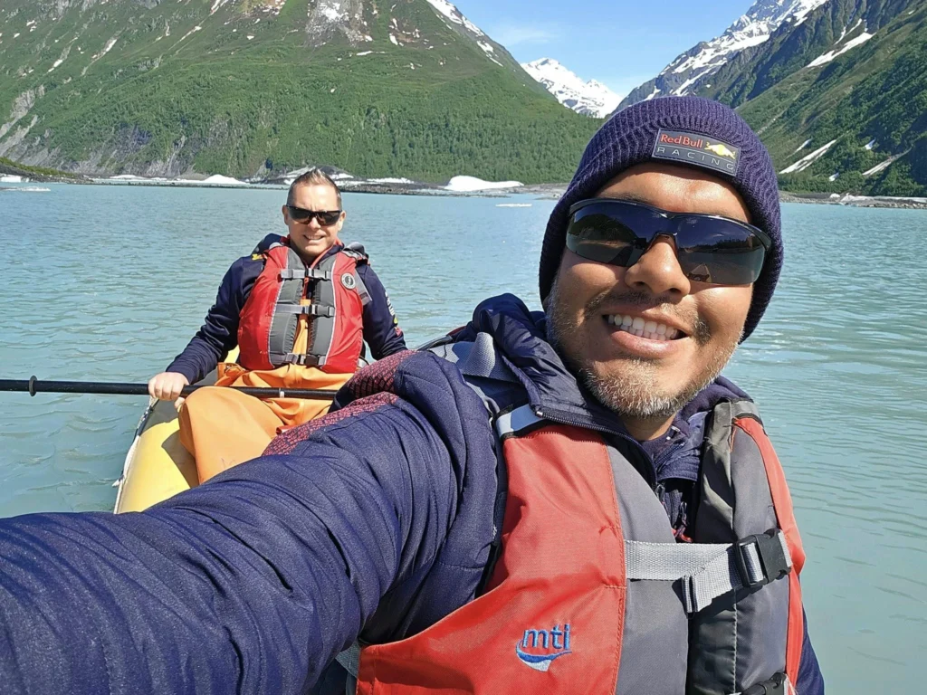 Cruising with Danny and Peter, Holland America Line, Westerdam, Prince William Sound, Valdez, Alaska, United States | A serene panorama of Prince William Sound at dawn, with the luxurious Westerdam cruise ship gliding through calm waters, surrounded by snow-capped mountains and glowing glaciers under a soft pink sky. Humpback whales breach the surface, their tails splashing against the reflective water, while sea otters float playfully in kelp beds and a bald eagle soars overhead. In Valdez Glacier Lake, vibrant blue icebergs drift peacefully, with kayakers in colorful kayaks paddling through the icy maze, dwarfed by the towering Valdez Glacier’s rugged ice face in the background. The Westerdam’s elegant deck features passengers enjoying a gourmet breakfast with panoramic views of the Alaskan wilderness, capturing the harmonious blend of luxury and nature.