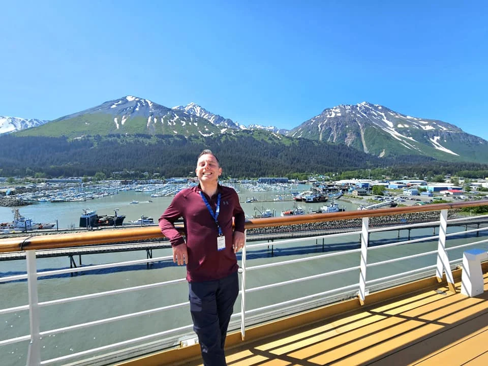 Cruising with Danny and Peter, Holland America Line, Westerdam, Seward, Alaska, United States | A panoramic view of Seward Harbor with rugged cliffs and serene waters, capturing Alaska's wilderness on a photography adventure. Travelers document vibrant wildflowers and snow-capped peaks in Chugach National Forest during an Alaska wilderness cruise. Historical remnants of the 1890 Gold Rush in Seward, including old mining equipment in lush alpine meadows, offer perfect scenes for photography enthusiasts. A majestic bald eagle soars over the tranquil waters of Seward Harbor, while senior-friendly guided tours explore accessible trails amidst stunning natural landscapes.
