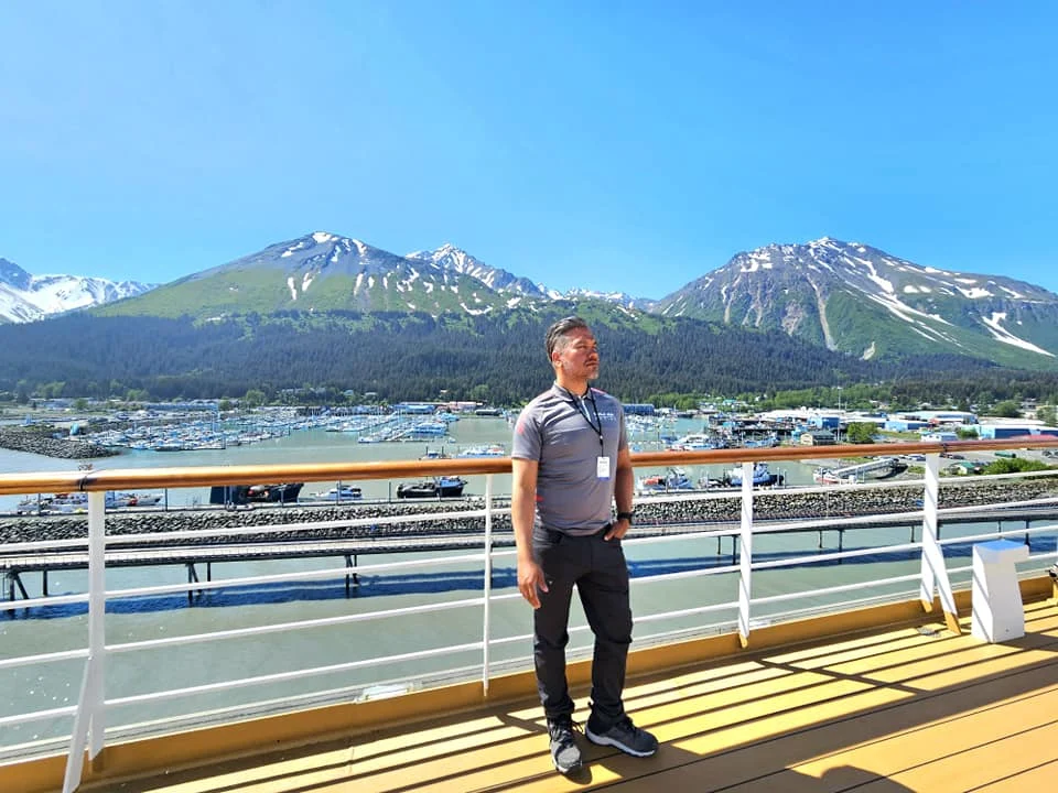 Cruising with Danny and Peter, Holland America Line, Westerdam, Seward, Alaska, United States | A panoramic view of Seward Harbor with rugged cliffs and serene waters, capturing Alaska's wilderness on a photography adventure. Travelers document vibrant wildflowers and snow-capped peaks in Chugach National Forest during an Alaska wilderness cruise. Historical remnants of the 1890 Gold Rush in Seward, including old mining equipment in lush alpine meadows, offer perfect scenes for photography enthusiasts. A majestic bald eagle soars over the tranquil waters of Seward Harbor, while senior-friendly guided tours explore accessible trails amidst stunning natural landscapes.