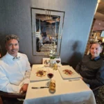 Cruising with Danny and Peter, Holland America Line, Westerdam, Bering Sea, Alaska, United States, Cellar Masters Dinner | A serene view of the Bering Sea from the deck of Holland America Line’s Westerdam during the Alaska Arctic Circle Solstice, with golden sunlight reflecting on calm, shimmering waters under a vast sky. In the distance, a pod of orcas glides gracefully, their black fins cutting through the sea. The ship’s elegant exterior is visible, with passengers wrapped in blankets, gazing at the horizon. Inside, the luxurious Pinnacle Grill dining room glows with soft lighting, showcasing a beautifully set table with crisp white linens, gleaming wine glasses, and a five-course Cellar Master’s Dinner. Plates feature duck confit with roasted cauliflower, porcini soup, warm lobster salad, wagyu beef with mushroom foam, and chocolate pot au crème with Grand Marnier. A sommelier pours Cloudy Bay Sauvignon Blanc, while guests savor the gourmet meal. The scene captures the harmony of Alaska’s wild beauty and refined culinary artistry, with the endless solstice daylight casting a warm, ethereal glow over the sea and ship, blending nature’s grandeur with sophisticated dining.