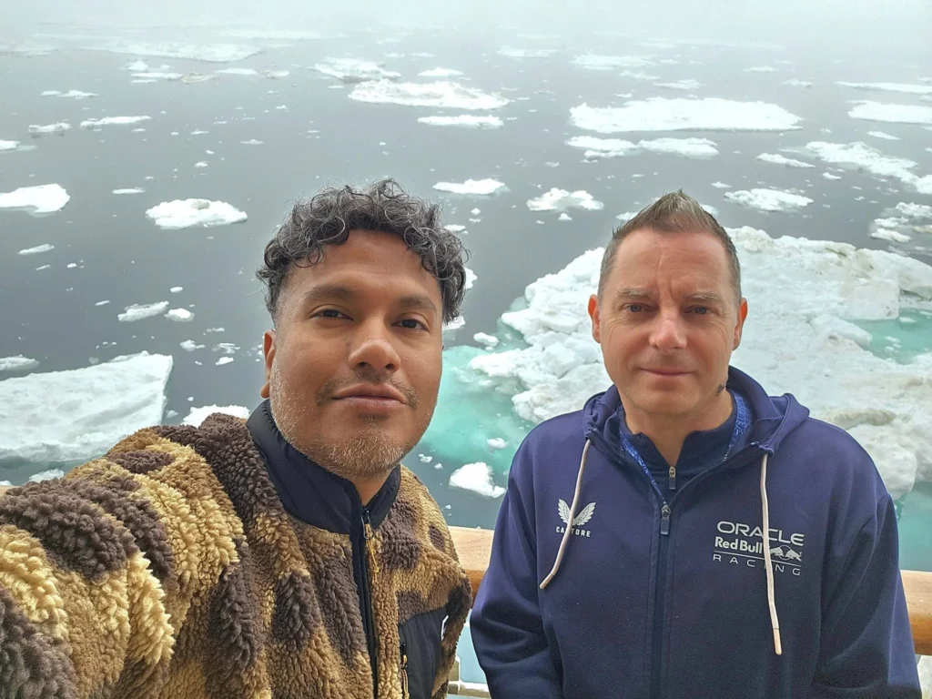 Cruising with Danny and Peter, Holland America Line, Westerdam, Bering Sea, Alaska, United States, Arctic Circle Adventure | A vibrant scene aboard Holland America Line’s Westerdam during the Alaska Arctic Circle Solstice cruise in the Bering Sea, Alaska, United States. Passengers gather around the Lido pool, preparing for the exhilarating Polar Bear Plunge, with crew members setting up the chilled pool to simulate Arctic waters. In the background, stunning Arctic landscapes unfold, featuring pristine ice floes floating on deep blue waters under a golden-hued sky, with distant glaciers and occasional wildlife, such as seals and seabirds, enhancing the untamed beauty of the Frozen Sea Adventure.