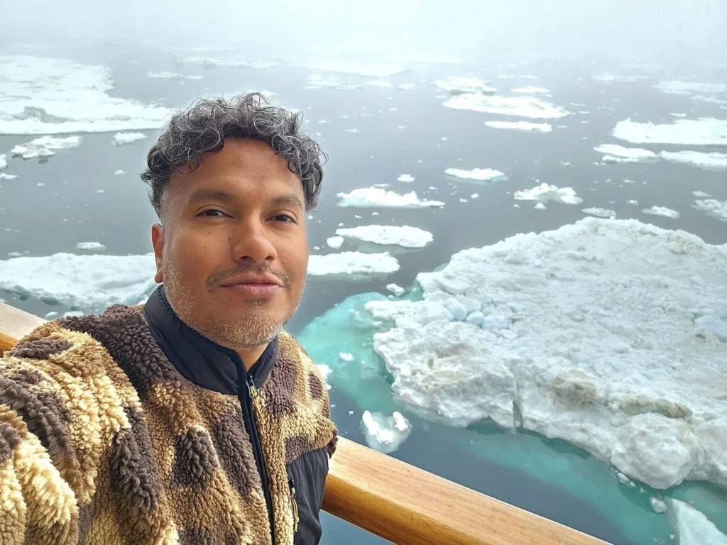 Cruising with Danny and Peter, Holland America Line, Westerdam, Bering Sea, Alaska, United States, Arctic Circle Adventure | A vibrant scene aboard Holland America Line’s Westerdam during the Alaska Arctic Circle Solstice cruise in the Bering Sea, Alaska, United States. Passengers gather around the Lido pool, preparing for the exhilarating Polar Bear Plunge, with crew members setting up the chilled pool to simulate Arctic waters. In the background, stunning Arctic landscapes unfold, featuring pristine ice floes floating on deep blue waters under a golden-hued sky, with distant glaciers and occasional wildlife, such as seals and seabirds, enhancing the untamed beauty of the Frozen Sea Adventure.