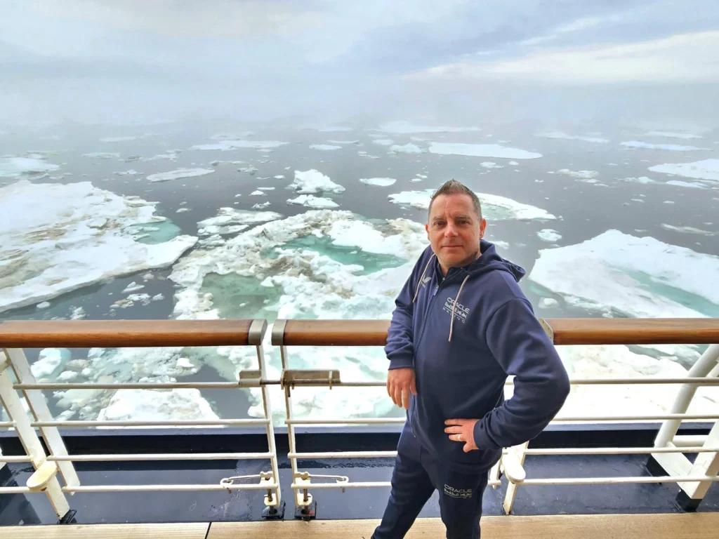 Cruising with Danny and Peter, Holland America Line, Westerdam, Bering Sea, Alaska, United States, Arctic Circle Adventure | A vibrant scene aboard Holland America Line’s Westerdam during the Alaska Arctic Circle Solstice cruise in the Bering Sea, Alaska, United States. Passengers gather around the Lido pool, preparing for the exhilarating Polar Bear Plunge, with crew members setting up the chilled pool to simulate Arctic waters. In the background, stunning Arctic landscapes unfold, featuring pristine ice floes floating on deep blue waters under a golden-hued sky, with distant glaciers and occasional wildlife, such as seals and seabirds, enhancing the untamed beauty of the Frozen Sea Adventure.