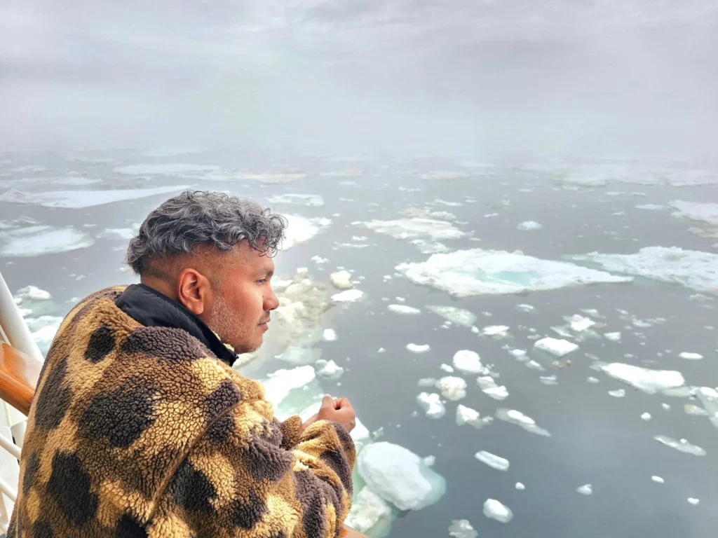 Cruising with Danny and Peter, Holland America Line, Westerdam, Bering Sea, Alaska, United States, Arctic Circle Adventure | A vibrant scene aboard Holland America Line’s Westerdam during the Alaska Arctic Circle Solstice cruise in the Bering Sea, Alaska, United States. Passengers gather around the Lido pool, preparing for the exhilarating Polar Bear Plunge, with crew members setting up the chilled pool to simulate Arctic waters. In the background, stunning Arctic landscapes unfold, featuring pristine ice floes floating on deep blue waters under a golden-hued sky, with distant glaciers and occasional wildlife, such as seals and seabirds, enhancing the untamed beauty of the Frozen Sea Adventure.