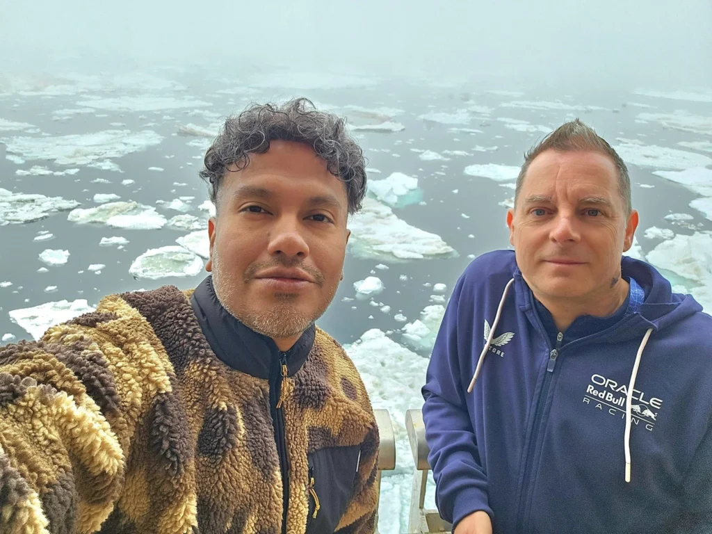 Cruising with Danny and Peter, Holland America Line, Westerdam, Bering Sea, Alaska, United States, Arctic Circle Adventure | A vibrant scene aboard Holland America Line’s Westerdam during the Alaska Arctic Circle Solstice cruise in the Bering Sea, Alaska, United States. Passengers gather around the Lido pool, preparing for the exhilarating Polar Bear Plunge, with crew members setting up the chilled pool to simulate Arctic waters. In the background, stunning Arctic landscapes unfold, featuring pristine ice floes floating on deep blue waters under a golden-hued sky, with distant glaciers and occasional wildlife, such as seals and seabirds, enhancing the untamed beauty of the Frozen Sea Adventure.