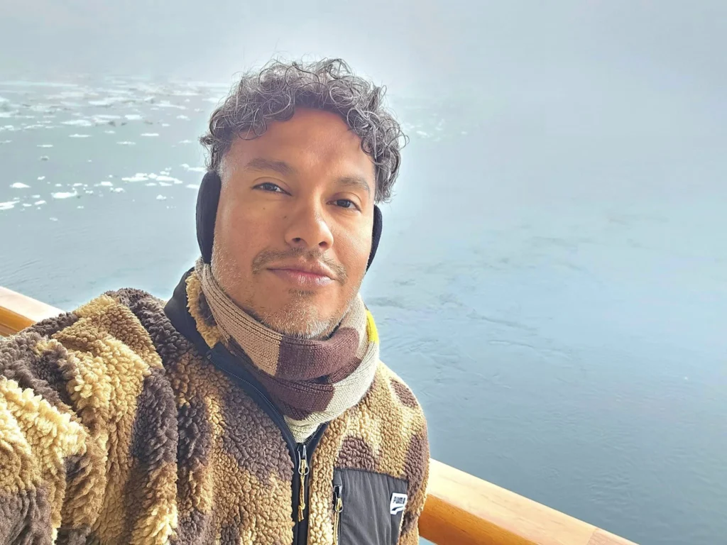 Cruising with Danny and Peter, Holland America Line, Westerdam, Bering Sea, Alaska, United States, Arctic Circle Adventure | A vibrant scene aboard Holland America Line’s Westerdam during the Alaska Arctic Circle Solstice cruise in the Bering Sea, Alaska, United States. Passengers gather around the Lido pool, preparing for the exhilarating Polar Bear Plunge, with crew members setting up the chilled pool to simulate Arctic waters. In the background, stunning Arctic landscapes unfold, featuring pristine ice floes floating on deep blue waters under a golden-hued sky, with distant glaciers and occasional wildlife, such as seals and seabirds, enhancing the untamed beauty of the Frozen Sea Adventure.