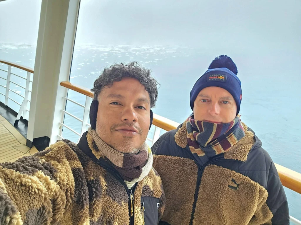 Cruising with Danny and Peter, Holland America Line, Westerdam, Bering Sea, Alaska, United States, Arctic Circle Adventure | A vibrant scene aboard Holland America Line’s Westerdam during the Alaska Arctic Circle Solstice cruise in the Bering Sea, Alaska, United States. Passengers gather around the Lido pool, preparing for the exhilarating Polar Bear Plunge, with crew members setting up the chilled pool to simulate Arctic waters. In the background, stunning Arctic landscapes unfold, featuring pristine ice floes floating on deep blue waters under a golden-hued sky, with distant glaciers and occasional wildlife, such as seals and seabirds, enhancing the untamed beauty of the Frozen Sea Adventure.