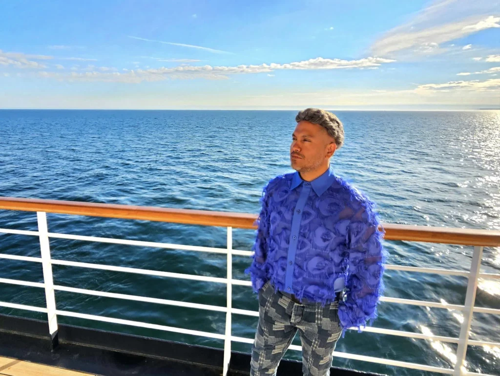 Cruising with Danny and Peter, Holland America Line, Westerdam, Bering Sea, Alaska, United States, Arctic Circle Adventure | A vibrant scene aboard Holland America Line’s Westerdam during the Alaska Arctic Circle Solstice cruise in the Bering Sea, Alaska, United States. Passengers gather around the Lido pool, preparing for the exhilarating Polar Bear Plunge, with crew members setting up the chilled pool to simulate Arctic waters. In the background, stunning Arctic landscapes unfold, featuring pristine ice floes floating on deep blue waters under a golden-hued sky, with distant glaciers and occasional wildlife, such as seals and seabirds, enhancing the untamed beauty of the Frozen Sea Adventure.