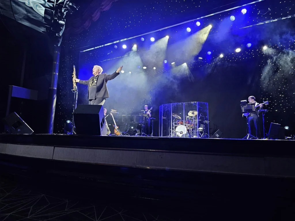 Cruising with Danny and Peter, Holland America Line, Westerdam, Bering Sea, Alaska, United States, Arctic Circle Adventure | A vibrant scene aboard Holland America Line’s Westerdam during the Alaska Arctic Circle Solstice cruise in the Bering Sea, Alaska, United States. Passengers gather around the Lido pool, preparing for the exhilarating Polar Bear Plunge, with crew members setting up the chilled pool to simulate Arctic waters. In the background, stunning Arctic landscapes unfold, featuring pristine ice floes floating on deep blue waters under a golden-hued sky, with distant glaciers and occasional wildlife, such as seals and seabirds, enhancing the untamed beauty of the Frozen Sea Adventure.