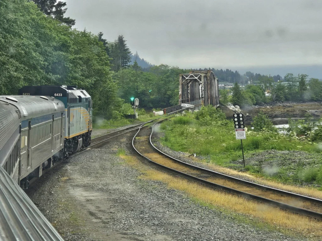 Cruising with Danny and Peter, Holland America Line, Westerdam, Prince Rupert, British Columbia, Canada | In the heart of British Columbia’s wild embrace, Where the Great Bear Rainforest weaves its mystic lace, A journey unfolds, both rail and road in stride, Through nature’s grandeur, where dreams and landscapes collide. From Prince Rupert’s charm, the train departs with grace, A serpentine path through forests’ ancient face, Panoramic windows frame a living, verdant dream, As the Skeena River’s flow whispers its serene theme. Mountains rise like giants, draped in emerald hues, While bald eagles soar, their shadows fleeting views, Black bears wander softly through the forest deep, And mountain goats on rugged cliffs silently leap. Kleanza Creek’s waterfalls cascade with a roar, Nature’s symphony on the rainforest’s floor, And Exchamsiks Park, a jewel of glacial light, Where shimmering pools reflect the day’s pure delight. From rail to road, the scenic drive unveils, A tapestry of mountains and verdant trails, Each turn a canvas painted with the wild’s own hand, A journey through a wonderland, vast and grand. Back in Prince Rupert, where history finds its place, The Museum’s treasures weave a cultural grace, Totem poles and masks tell tales of ancient lore, In this land where the past and present gently soar. Through the Great Bear Rainforest, we wandered wide, In rail and road’s embrace, where nature and culture collide, A journey of wonder, where memories are spun, In the heart of British Columbia, under the sun.