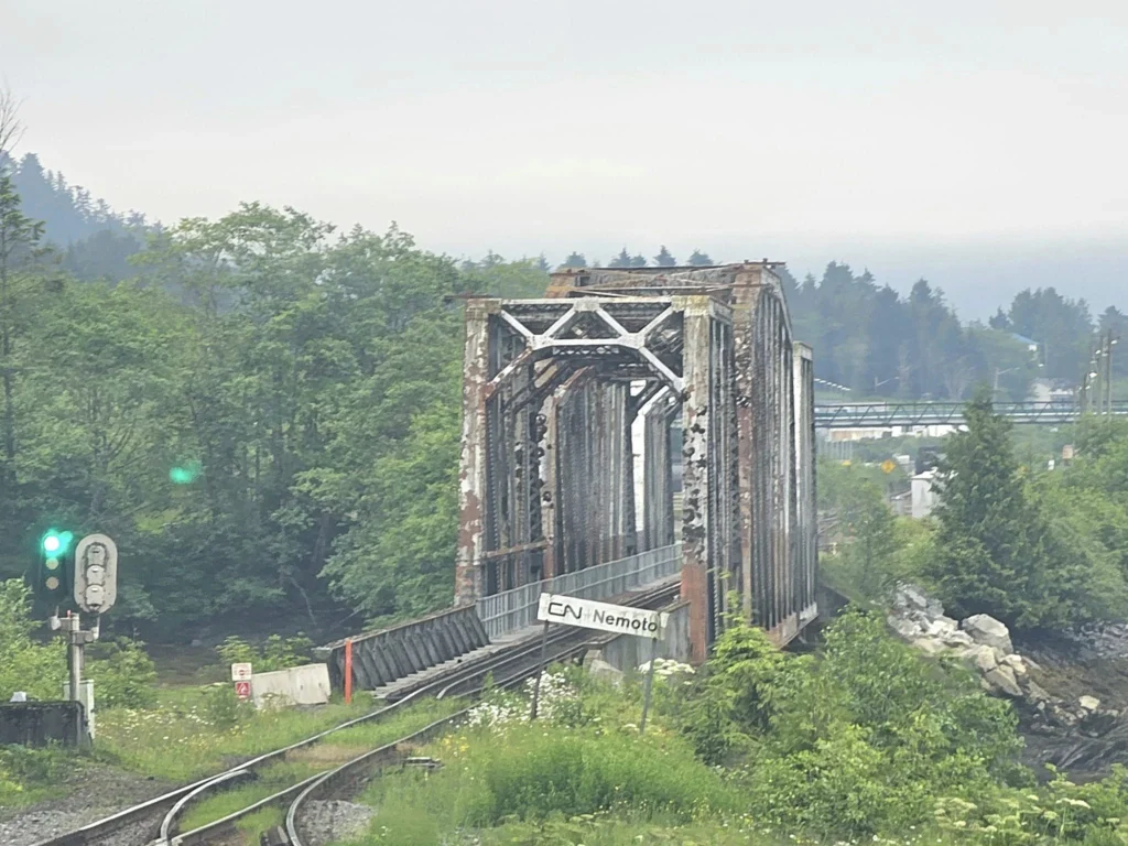 Cruising with Danny and Peter, Holland America Line, Westerdam, Prince Rupert, British Columbia, Canada | In the heart of British Columbia’s wild embrace, Where the Great Bear Rainforest weaves its mystic lace, A journey unfolds, both rail and road in stride, Through nature’s grandeur, where dreams and landscapes collide. From Prince Rupert’s charm, the train departs with grace, A serpentine path through forests’ ancient face, Panoramic windows frame a living, verdant dream, As the Skeena River’s flow whispers its serene theme. Mountains rise like giants, draped in emerald hues, While bald eagles soar, their shadows fleeting views, Black bears wander softly through the forest deep, And mountain goats on rugged cliffs silently leap. Kleanza Creek’s waterfalls cascade with a roar, Nature’s symphony on the rainforest’s floor, And Exchamsiks Park, a jewel of glacial light, Where shimmering pools reflect the day’s pure delight. From rail to road, the scenic drive unveils, A tapestry of mountains and verdant trails, Each turn a canvas painted with the wild’s own hand, A journey through a wonderland, vast and grand. Back in Prince Rupert, where history finds its place, The Museum’s treasures weave a cultural grace, Totem poles and masks tell tales of ancient lore, In this land where the past and present gently soar. Through the Great Bear Rainforest, we wandered wide, In rail and road’s embrace, where nature and culture collide, A journey of wonder, where memories are spun, In the heart of British Columbia, under the sun.