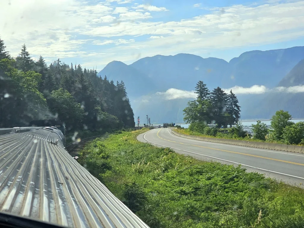 Cruising with Danny and Peter, Holland America Line, Westerdam, Prince Rupert, British Columbia, Canada | In the heart of British Columbia’s wild embrace, Where the Great Bear Rainforest weaves its mystic lace, A journey unfolds, both rail and road in stride, Through nature’s grandeur, where dreams and landscapes collide. From Prince Rupert’s charm, the train departs with grace, A serpentine path through forests’ ancient face, Panoramic windows frame a living, verdant dream, As the Skeena River’s flow whispers its serene theme. Mountains rise like giants, draped in emerald hues, While bald eagles soar, their shadows fleeting views, Black bears wander softly through the forest deep, And mountain goats on rugged cliffs silently leap. Kleanza Creek’s waterfalls cascade with a roar, Nature’s symphony on the rainforest’s floor, And Exchamsiks Park, a jewel of glacial light, Where shimmering pools reflect the day’s pure delight. From rail to road, the scenic drive unveils, A tapestry of mountains and verdant trails, Each turn a canvas painted with the wild’s own hand, A journey through a wonderland, vast and grand. Back in Prince Rupert, where history finds its place, The Museum’s treasures weave a cultural grace, Totem poles and masks tell tales of ancient lore, In this land where the past and present gently soar. Through the Great Bear Rainforest, we wandered wide, In rail and road’s embrace, where nature and culture collide, A journey of wonder, where memories are spun, In the heart of British Columbia, under the sun.