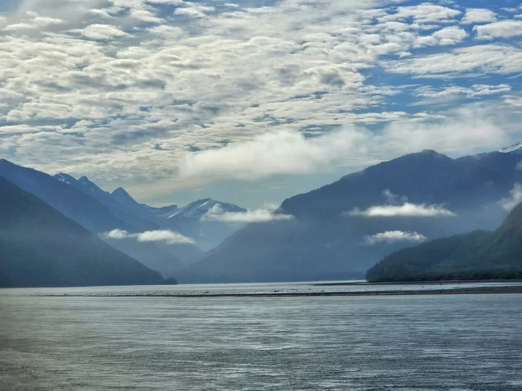 Cruising with Danny and Peter, Holland America Line, Westerdam, Prince Rupert, British Columbia, Canada | In the heart of British Columbia’s wild embrace, Where the Great Bear Rainforest weaves its mystic lace, A journey unfolds, both rail and road in stride, Through nature’s grandeur, where dreams and landscapes collide. From Prince Rupert’s charm, the train departs with grace, A serpentine path through forests’ ancient face, Panoramic windows frame a living, verdant dream, As the Skeena River’s flow whispers its serene theme. Mountains rise like giants, draped in emerald hues, While bald eagles soar, their shadows fleeting views, Black bears wander softly through the forest deep, And mountain goats on rugged cliffs silently leap. Kleanza Creek’s waterfalls cascade with a roar, Nature’s symphony on the rainforest’s floor, And Exchamsiks Park, a jewel of glacial light, Where shimmering pools reflect the day’s pure delight. From rail to road, the scenic drive unveils, A tapestry of mountains and verdant trails, Each turn a canvas painted with the wild’s own hand, A journey through a wonderland, vast and grand. Back in Prince Rupert, where history finds its place, The Museum’s treasures weave a cultural grace, Totem poles and masks tell tales of ancient lore, In this land where the past and present gently soar. Through the Great Bear Rainforest, we wandered wide, In rail and road’s embrace, where nature and culture collide, A journey of wonder, where memories are spun, In the heart of British Columbia, under the sun.