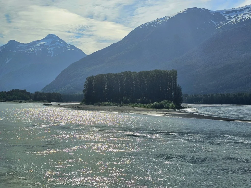 Cruising with Danny and Peter, Holland America Line, Westerdam, Prince Rupert, British Columbia, Canada | In the heart of British Columbia’s wild embrace, Where the Great Bear Rainforest weaves its mystic lace, A journey unfolds, both rail and road in stride, Through nature’s grandeur, where dreams and landscapes collide. From Prince Rupert’s charm, the train departs with grace, A serpentine path through forests’ ancient face, Panoramic windows frame a living, verdant dream, As the Skeena River’s flow whispers its serene theme. Mountains rise like giants, draped in emerald hues, While bald eagles soar, their shadows fleeting views, Black bears wander softly through the forest deep, And mountain goats on rugged cliffs silently leap. Kleanza Creek’s waterfalls cascade with a roar, Nature’s symphony on the rainforest’s floor, And Exchamsiks Park, a jewel of glacial light, Where shimmering pools reflect the day’s pure delight. From rail to road, the scenic drive unveils, A tapestry of mountains and verdant trails, Each turn a canvas painted with the wild’s own hand, A journey through a wonderland, vast and grand. Back in Prince Rupert, where history finds its place, The Museum’s treasures weave a cultural grace, Totem poles and masks tell tales of ancient lore, In this land where the past and present gently soar. Through the Great Bear Rainforest, we wandered wide, In rail and road’s embrace, where nature and culture collide, A journey of wonder, where memories are spun, In the heart of British Columbia, under the sun.