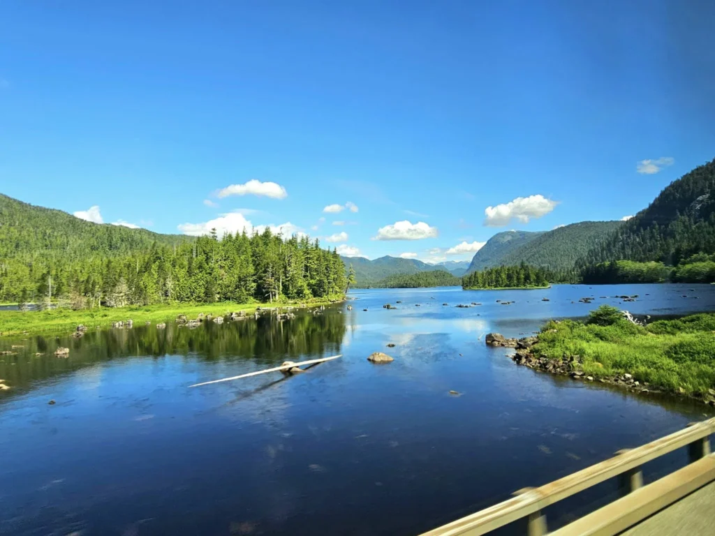 Cruising with Danny and Peter, Holland America Line, Westerdam, Prince Rupert, British Columbia, Canada | In the heart of British Columbia’s wild embrace, Where the Great Bear Rainforest weaves its mystic lace, A journey unfolds, both rail and road in stride, Through nature’s grandeur, where dreams and landscapes collide. From Prince Rupert’s charm, the train departs with grace, A serpentine path through forests’ ancient face, Panoramic windows frame a living, verdant dream, As the Skeena River’s flow whispers its serene theme. Mountains rise like giants, draped in emerald hues, While bald eagles soar, their shadows fleeting views, Black bears wander softly through the forest deep, And mountain goats on rugged cliffs silently leap. Kleanza Creek’s waterfalls cascade with a roar, Nature’s symphony on the rainforest’s floor, And Exchamsiks Park, a jewel of glacial light, Where shimmering pools reflect the day’s pure delight. From rail to road, the scenic drive unveils, A tapestry of mountains and verdant trails, Each turn a canvas painted with the wild’s own hand, A journey through a wonderland, vast and grand. Back in Prince Rupert, where history finds its place, The Museum’s treasures weave a cultural grace, Totem poles and masks tell tales of ancient lore, In this land where the past and present gently soar. Through the Great Bear Rainforest, we wandered wide, In rail and road’s embrace, where nature and culture collide, A journey of wonder, where memories are spun, In the heart of British Columbia, under the sun.