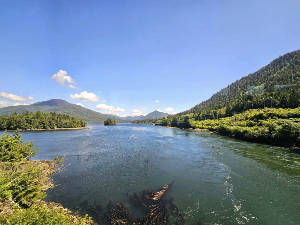 Cruising with Danny and Peter, Holland America Line, Westerdam, Prince Rupert, British Columbia, Canada | In the heart of British Columbia’s wild embrace, Where the Great Bear Rainforest weaves its mystic lace, A journey unfolds, both rail and road in stride, Through nature’s grandeur, where dreams and landscapes collide. From Prince Rupert’s charm, the train departs with grace, A serpentine path through forests’ ancient face, Panoramic windows frame a living, verdant dream, As the Skeena River’s flow whispers its serene theme. Mountains rise like giants, draped in emerald hues, While bald eagles soar, their shadows fleeting views, Black bears wander softly through the forest deep, And mountain goats on rugged cliffs silently leap. Kleanza Creek’s waterfalls cascade with a roar, Nature’s symphony on the rainforest’s floor, And Exchamsiks Park, a jewel of glacial light, Where shimmering pools reflect the day’s pure delight. From rail to road, the scenic drive unveils, A tapestry of mountains and verdant trails, Each turn a canvas painted with the wild’s own hand, A journey through a wonderland, vast and grand. Back in Prince Rupert, where history finds its place, The Museum’s treasures weave a cultural grace, Totem poles and masks tell tales of ancient lore, In this land where the past and present gently soar. Through the Great Bear Rainforest, we wandered wide, In rail and road’s embrace, where nature and culture collide, A journey of wonder, where memories are spun, In the heart of British Columbia, under the sun.
