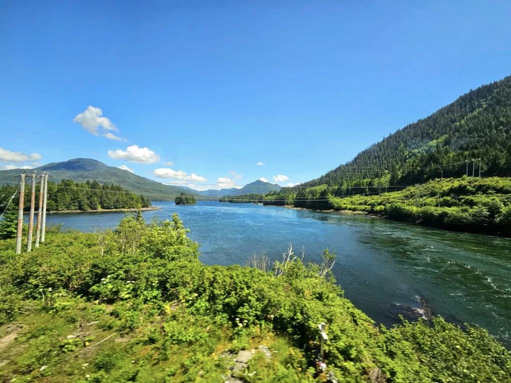 Cruising with Danny and Peter, Holland America Line, Westerdam, Prince Rupert, British Columbia, Canada | In the heart of British Columbia’s wild embrace, Where the Great Bear Rainforest weaves its mystic lace, A journey unfolds, both rail and road in stride, Through nature’s grandeur, where dreams and landscapes collide. From Prince Rupert’s charm, the train departs with grace, A serpentine path through forests’ ancient face, Panoramic windows frame a living, verdant dream, As the Skeena River’s flow whispers its serene theme. Mountains rise like giants, draped in emerald hues, While bald eagles soar, their shadows fleeting views, Black bears wander softly through the forest deep, And mountain goats on rugged cliffs silently leap. Kleanza Creek’s waterfalls cascade with a roar, Nature’s symphony on the rainforest’s floor, And Exchamsiks Park, a jewel of glacial light, Where shimmering pools reflect the day’s pure delight. From rail to road, the scenic drive unveils, A tapestry of mountains and verdant trails, Each turn a canvas painted with the wild’s own hand, A journey through a wonderland, vast and grand. Back in Prince Rupert, where history finds its place, The Museum’s treasures weave a cultural grace, Totem poles and masks tell tales of ancient lore, In this land where the past and present gently soar. Through the Great Bear Rainforest, we wandered wide, In rail and road’s embrace, where nature and culture collide, A journey of wonder, where memories are spun, In the heart of British Columbia, under the sun.