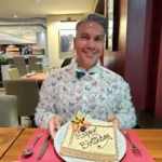 Cruising with Danny and Peter, MSC Cruises, MSC Preziosa, Rotterdam, The Netherlands, Birthday Cruise | The majestic MSC Preziosa cruise ship docked at Rotterdam’s Cruise Terminal on Wilhelminakade, its sleek white exterior gleaming against the modern city skyline with the iconic Erasmus Bridge in the background. Passengers board for a Northern Europe mini-cruise, celebrating a birthday with luxurious touches like a chilled bottle of Spumante and macarons in a stylish cabin. The vibrant onboard atmosphere features a gourmet dining experience at Butcher’s Cut Chef’s Table with a surprise birthday cake, thrilling entertainment at the Platinum Theater’s Rock Nation show with singers and dancers, and a lively Silent Disco in the Safari Lounge. The scene captures the excitement of embarkation day, with scenic views of Rotterdam’s waterfront, onboard shopping for MSC souvenirs, and the promise of an unforgettable luxury cruise vacation.